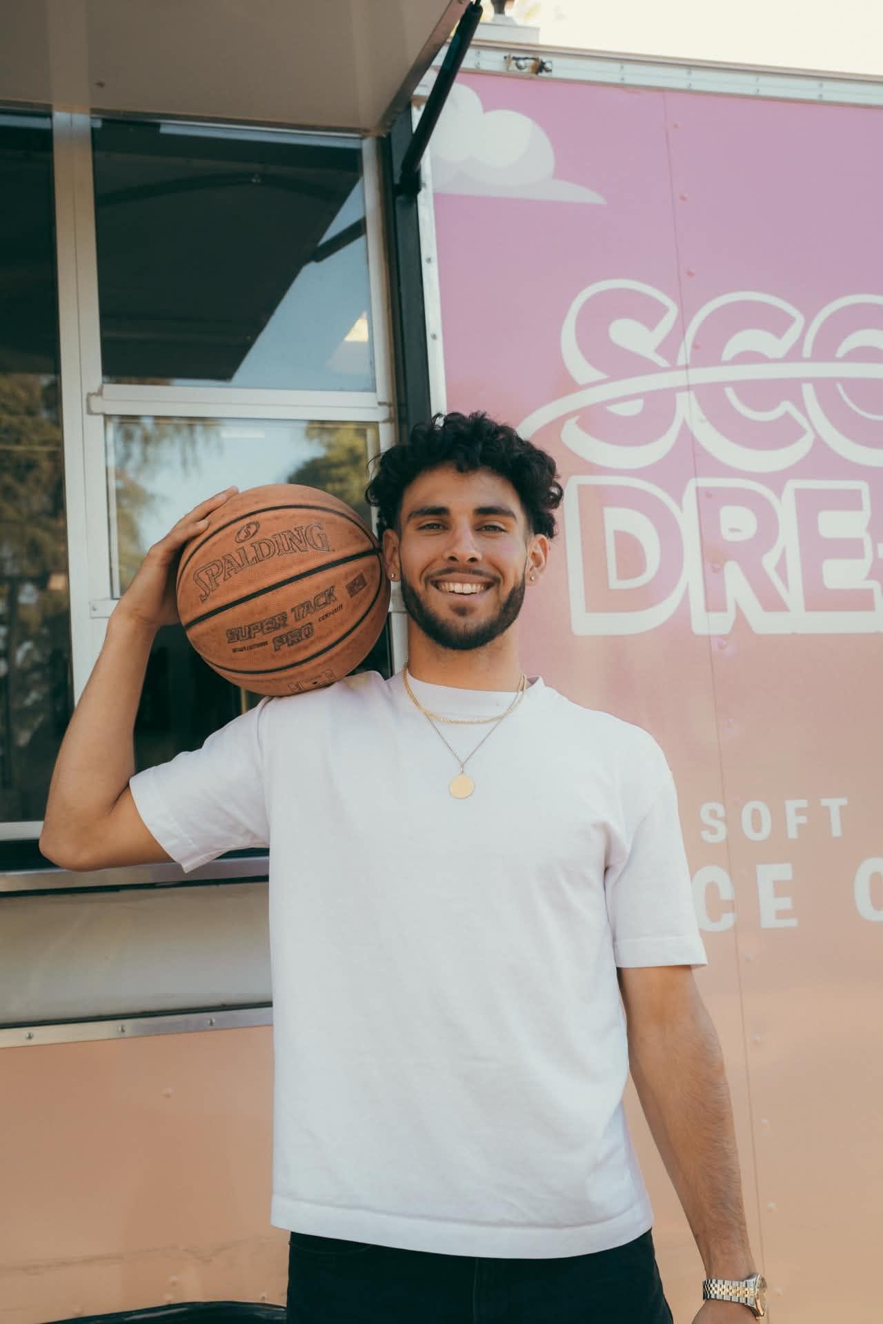 Devan Minouei smiling with a Spalding basketball over his shoulder in front of the Scoop Dreams trailer