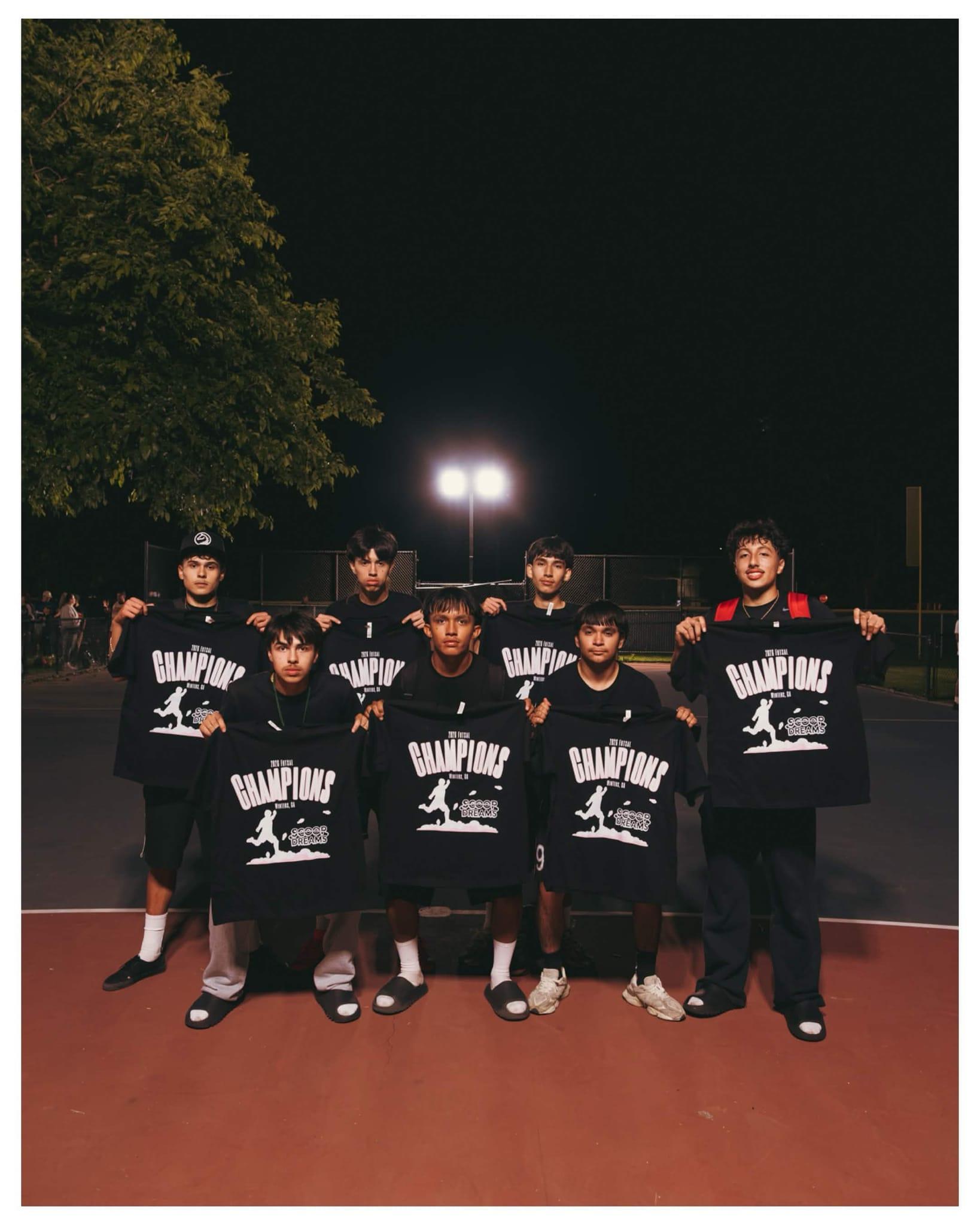 Championship team holding their Scoop Dreams 3-on-3 tournament shirts on the court at night