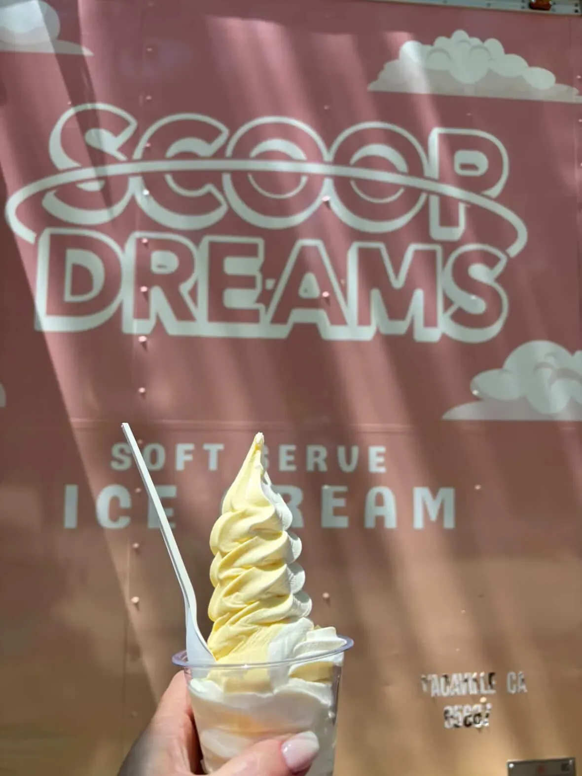 A cup of swirled vanilla soft serve held up in front of the pink Scoop Dreams trailer reading Soft Serve Ice Cream, Vacaville CA